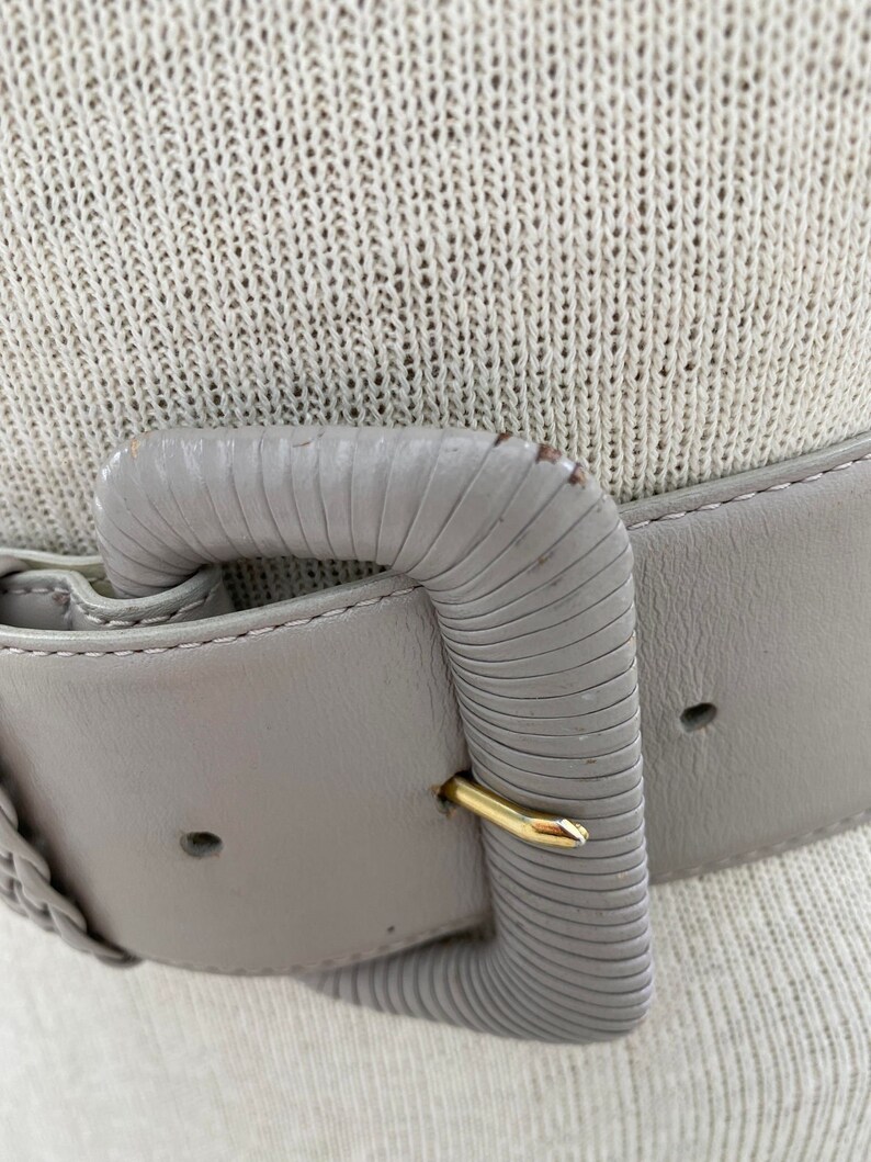 Puede incluir: Un cintur&oacute;n de cuero gris con una hebilla texturizada. La hebilla es de forma rectangular simple con un cierre dorado.