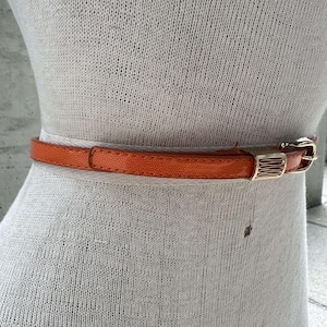 May include: A bright orange leather belt with a gold buckle. The belt is on a white mannequin.