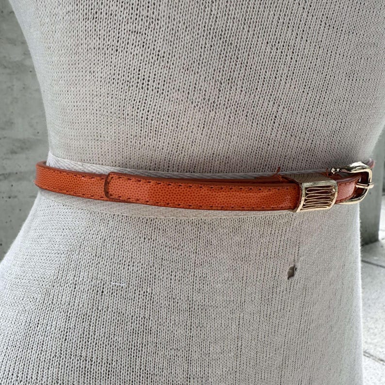 May include: A bright orange leather belt with a gold buckle. The belt is on a white mannequin.