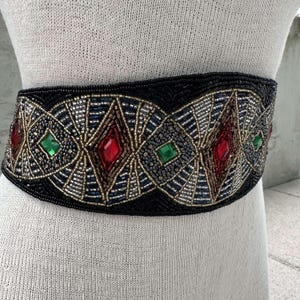 Pu&ograve; includere: Una cintura nera di perline con un design geometrico con accenti rossi, verdi e dorati. La cintura &egrave; adornata con grandi pietre preziose sfaccettate.