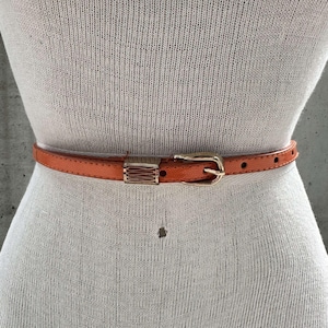 May include: A bright orange leather belt with a gold buckle. The belt is on a white mannequin.