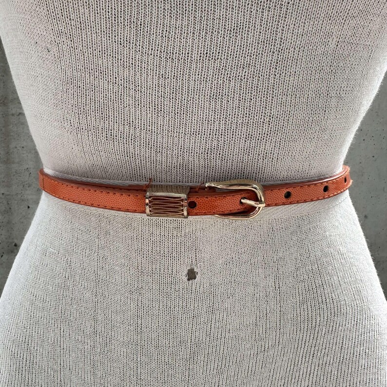 May include: A bright orange leather belt with a gold buckle. The belt is on a white mannequin.