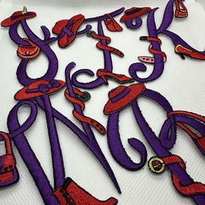 Peut inclure: Lettres brodées en violet orthographiant "New York" avec des accents rouges de chapeaux, de chaussures et de sacs à main.