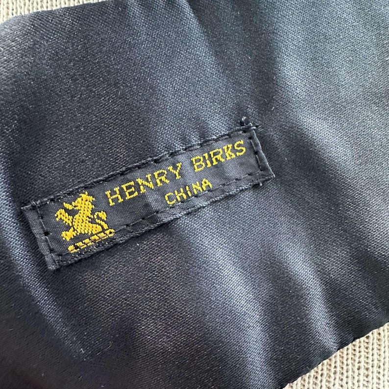 Pu&ograve; includere: Un'etichetta in tessuto nero con testo dorato che recita "HENRY BIRKS CHINA" e un logo di leone dorato.