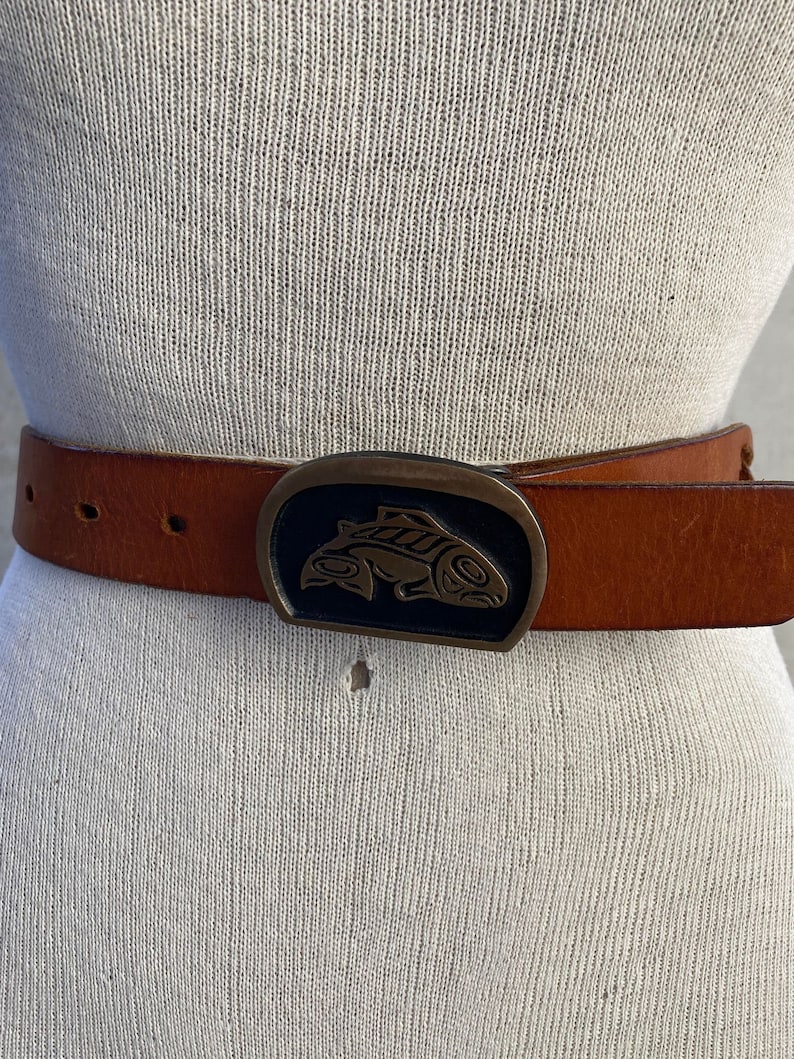 May include: A brown leather belt with a large, oval-shaped buckle featuring a stylized fish design in black and gold.