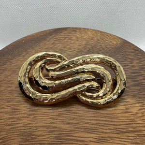 Może przedstawiać: Złota, fakturowana broszka z podwójną pętlą w kształcie ósemki.