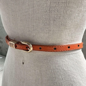 May include: A close-up of a brown leather belt with a gold buckle. The belt is on a white knitted fabric background.