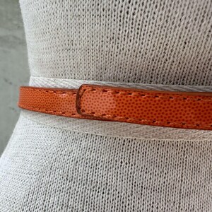 May include: A close-up of a bright orange leather belt with a textured surface. The belt is on a white fabric background.