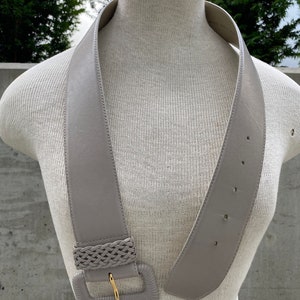 Puede incluir: Un cintur&oacute;n ancho de cuero gris con una hebilla trenzada. La hebilla es de forma rectangular y tiene un cierre de metal dorado.