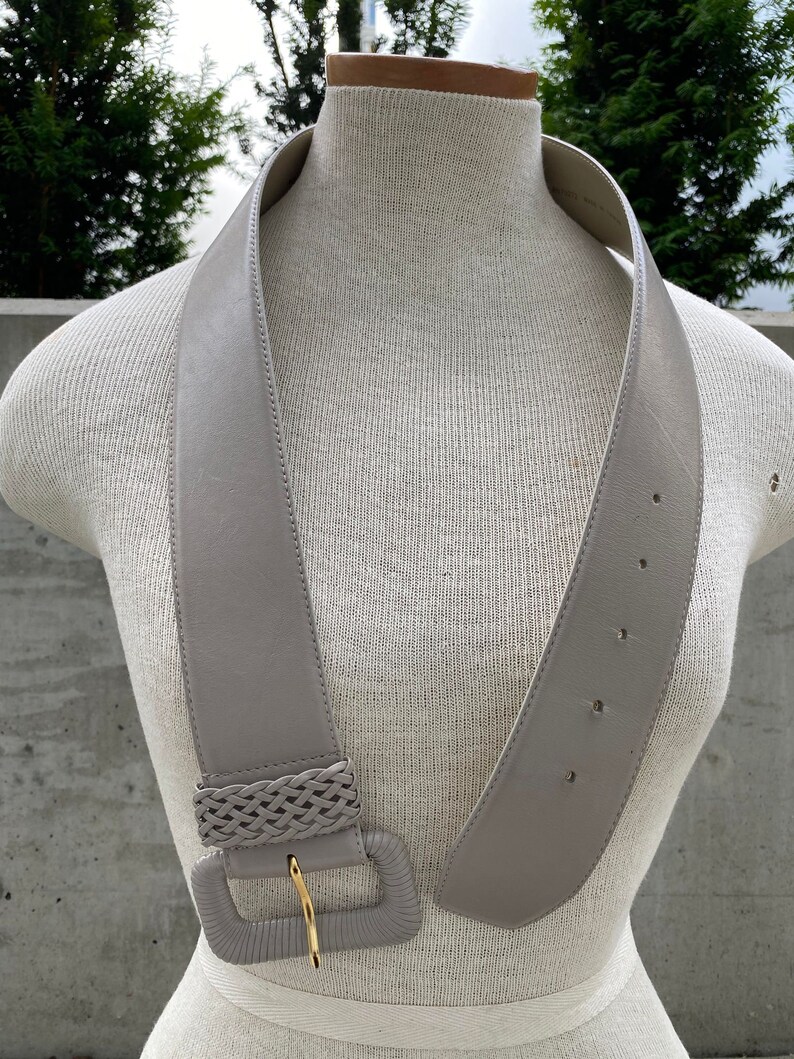 Puede incluir: Un cintur&oacute;n ancho de cuero gris con una hebilla trenzada. La hebilla es de forma rectangular y tiene un cierre de metal dorado.
