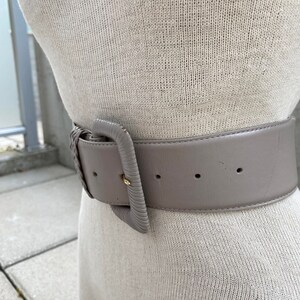Puede incluir: Un cintur&oacute;n ancho de cuero gris con una hebilla grande y rectangular. La hebilla es de metal claro y tiene una superficie ligeramente texturizada.