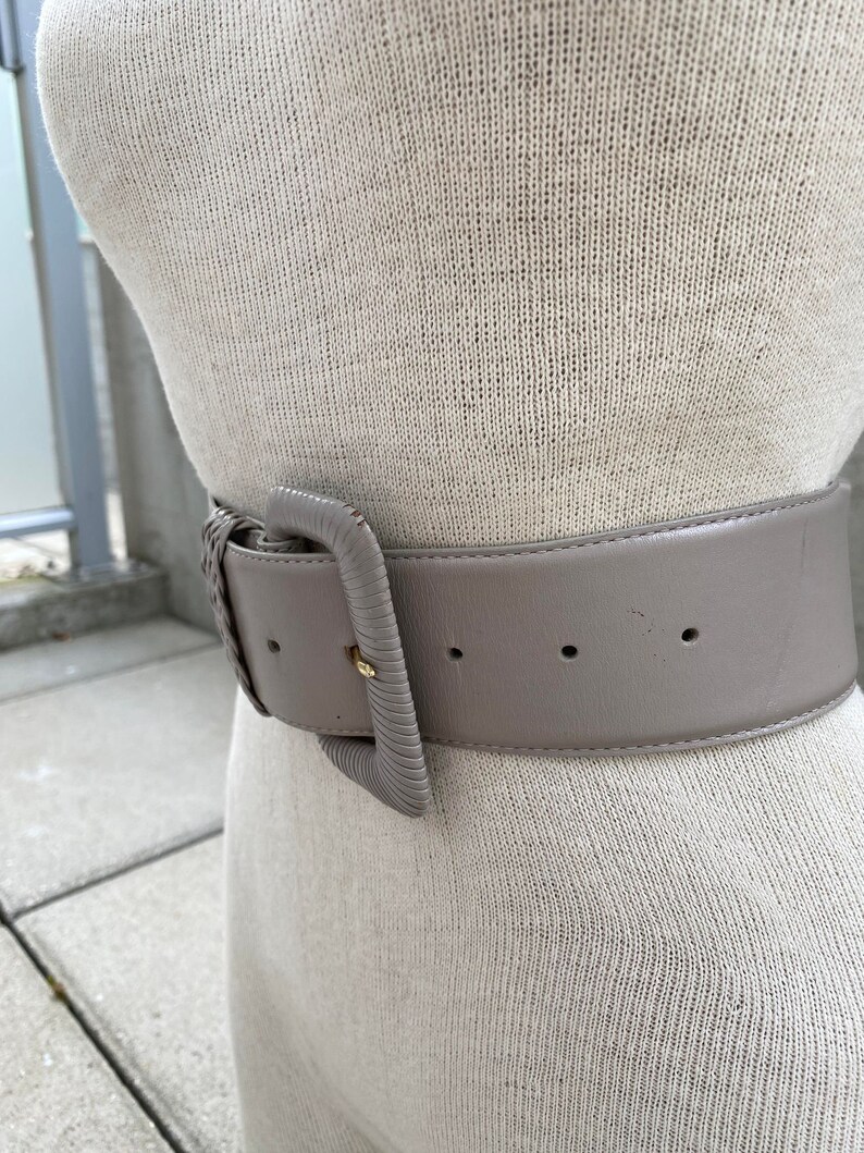 Puede incluir: Un cintur&oacute;n ancho de cuero gris con una hebilla grande y rectangular. La hebilla es de metal claro y tiene una superficie ligeramente texturizada.