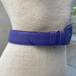 May include: A vibrant purple belt with a textured, lizard-skin-like pattern and a matching purple buckle. The belt features two loops and a pointed tip, secured around a beige mannequin torso to display its fit and color.