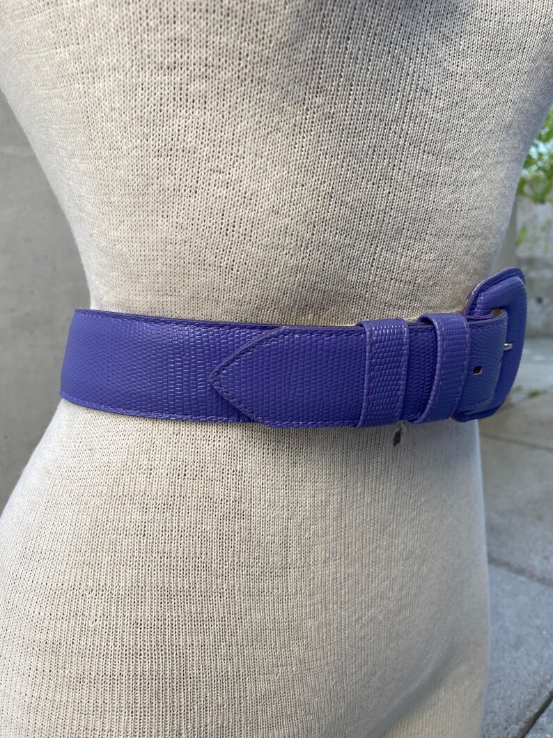 May include: A vibrant purple belt with a textured, lizard-skin-like pattern and a matching purple buckle. The belt features two loops and a pointed tip, secured around a beige mannequin torso to display its fit and color.