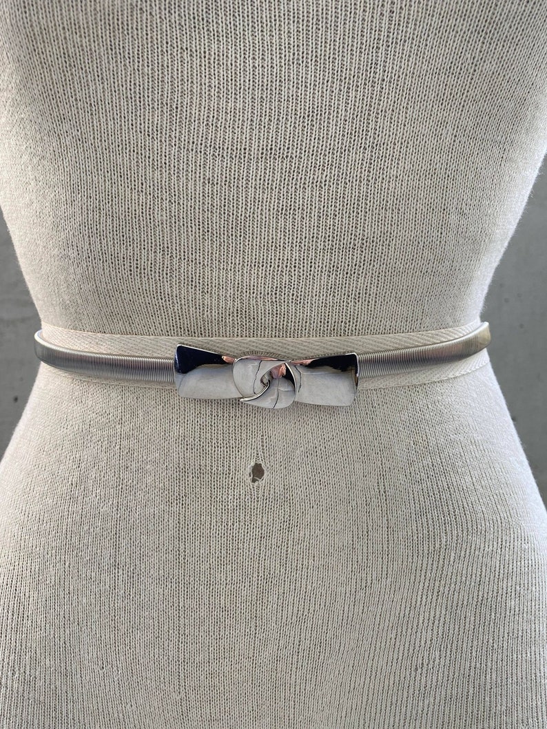 May include: A silver metal belt with a bow-shaped buckle. The belt is on a white mannequin.