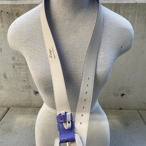 May include: Cream-colored leather belt with a purple and cream buckle. The belt has a purple lining and is displayed on a mannequin. The belt is labeled "Large" and "Made in Canada".