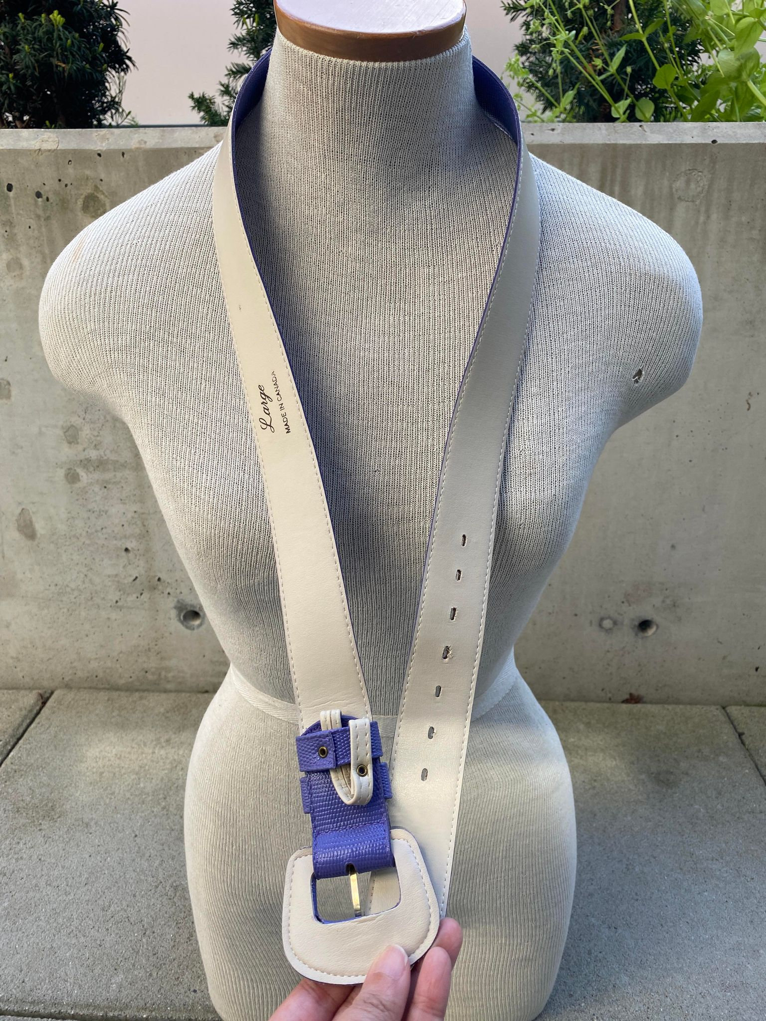 May include: Cream-colored leather belt with a purple and cream buckle. The belt has a purple lining and is displayed on a mannequin. The belt is labeled "Large" and "Made in Canada".