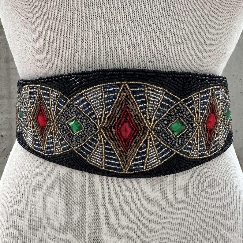Pu&ograve; includere: Una cintura nera di perline con un design geometrico con accenti rossi, verdi e dorati. La cintura &egrave; adornata con grandi pietre preziose sfaccettate.