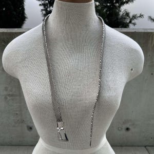 Peut inclure: Un collier argent&eacute; avec un motif en maille et une boucle rectangulaire. Le collier est pr&eacute;sent&eacute; sur un mannequin blanc. C'est un long accessoire &eacute;l&eacute;gant.