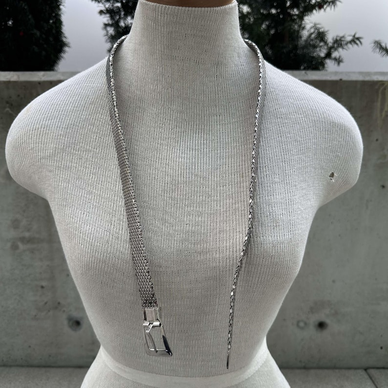 Peut inclure: Un collier argent&eacute; avec un motif en maille et une boucle rectangulaire. Le collier est pr&eacute;sent&eacute; sur un mannequin blanc. C'est un long accessoire &eacute;l&eacute;gant.