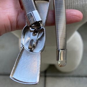 May include: A silver metal belt with a bow-shaped buckle. The buckle is closed and the belt is coiled.