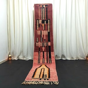 May include: A long, narrow rug with a geometric pattern in shades of pink, orange, black, and white. The rug has a fringe on both ends.