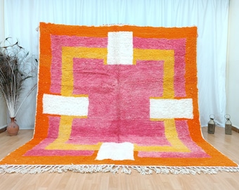 May include: A large, rectangular rug with a geometric design. The rug features a vibrant color palette of orange, pink, yellow, and white. The design includes a central pink field with white and yellow geometric shapes, framed by an orange border and fringe.
