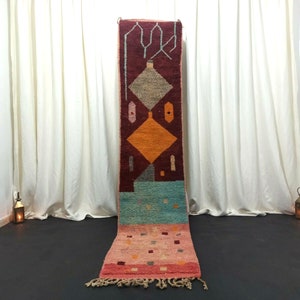 May include: A long, narrow, red, orange, blue, and pink Moroccan rug with a geometric pattern. The rug is hanging on a white wall in a room with white curtains.
