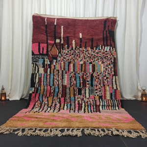 Peut inclure: Un tapis marocain coloré avec des motifs géométriques dans des tons de rose, bleu, marron et noir. Le tapis a une bordure à franges et est tissé avec une variété de textures.
