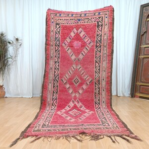 May include: A large, rectangular, red rug with a geometric diamond pattern. The rug has a dark brown border and fringe. The design includes diamond shapes and a central floral motif. The rug is displayed on a light wood floor.