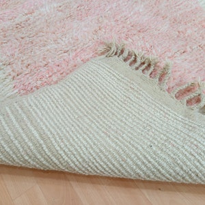 May include: A pink and white fluffy rug with a fringe border. The rug is folded in half, showing the underside, which is a woven fabric with a striped pattern.