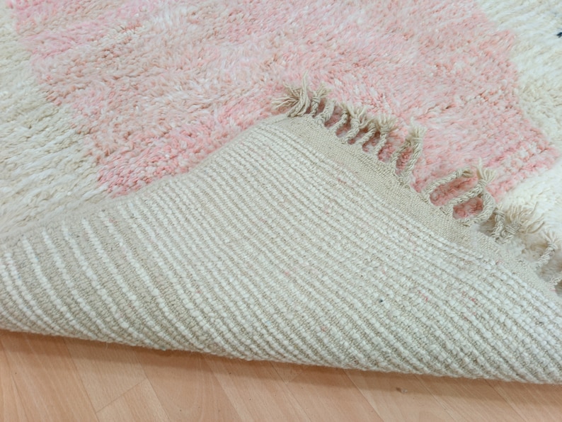 May include: A pink and white fluffy rug with a fringe border. The rug is folded in half, showing the underside, which is a woven fabric with a striped pattern.
