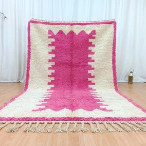 May include: A large, rectangular rug with a central pink design and a cream-colored border. The pink section features a geometric pattern. The rug has a fringe along the bottom edge and is displayed on a wooden floor.