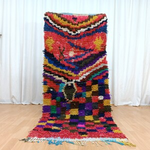 Könnte beinhalten: Ein bunter marokkanischer Teppich mit einem geometrischen Muster in Rot-, Blau-, Gelb-, Grün- und Violetttönen. Der Teppich hat einen Fransenrand und ist aus Wolle gefertigt.