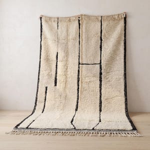 May include: A rectangular area rug featuring a thick, cream-colored wool pile with a series of bold, black vertical and horizontal lines forming an abstract geometric pattern. The rug is finished with knotted fringe along both short ends.