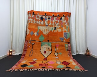 May include: A large rectangular area rug featuring a vibrant orange background with a variety of geometric shapes and abstract symbols in black, white, pink, green, and blue. The rug has a fringed edge and a repeating diamond pattern at the top.