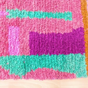 K&ouml;nnte beinhalten: Ein bunter, handgewebter Teppich mit einem geometrischen Muster in Rosat&ouml;nen, Lila, Gr&uuml;n und Braun. Der Teppich hat einen Fransenrand.