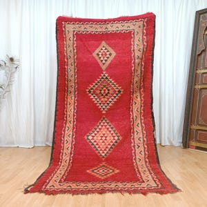 May include: A striking red rug featuring a geometric diamond design in black, tan, and orange. The rectangular rug has a decorative border. It is displayed against a white curtain backdrop.