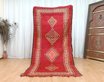 May include: A vibrant red rug with a geometric diamond pattern in black, tan, and orange. The rug has a rectangular shape and a decorative border. The rug is displayed against a white curtain backdrop.
