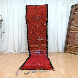 May include: A long, rectangular red rug with a black border and diamond pattern. The rug has a fringe at the bottom and is displayed on a wooden floor. The rug is a decorative floor covering.