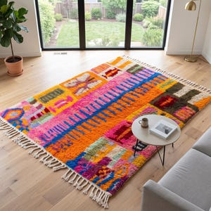 Op de afbeelding: Een rechthoekig vloerkleed met een levendig, veelkleurig geometrisch patroon met blokken van fel oranje, roze, geel, blauw en groen. Het kleed heeft een dikke, getextureerde pool en is aan beide uiteinden afgewerkt met crèmekleurige franjes.
