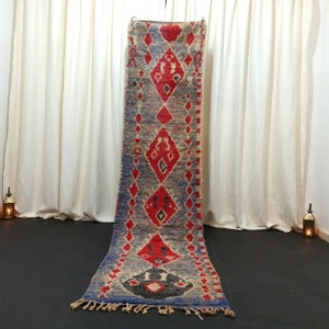 May include: A long, narrow rug with a geometric pattern in shades of red, blue, and white. The rug is hanging on a white wall with white curtains on either side.