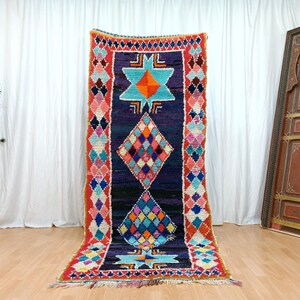 Op de afbeelding: Een rechthoekig, handgeweven tapijt met een donkerblauwe achtergrond en kleurrijke geometrische patronen. Het tapijt heeft ruit- en stervormen in tinten oranje, blauw, roze en groen, met een rode rand. Het tapijt wordt op een houten vloer getoond.