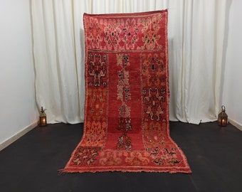 May include: A red and black Moroccan rug with intricate geometric patterns. The rug is laid out on a dark floor in front of a white curtain.