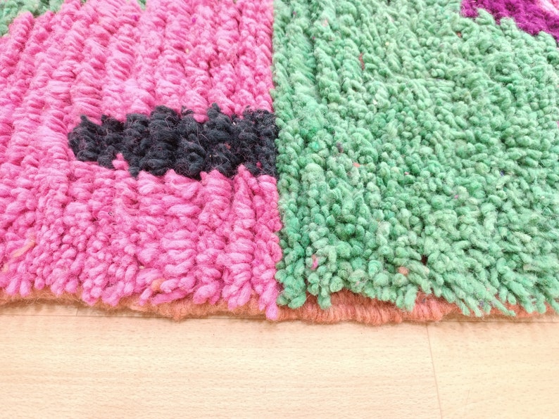 K&ouml;nnte beinhalten: Nahaufnahme eines rosa, gr&uuml;nen und schwarzen Teppichs mit einer flauschigen Textur. Der Teppich hat einen gewebten Rand.