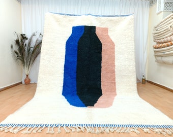 May include: A white wool rug with a geometric design featuring three vertical stripes in blue, black, and pink. The rug has a fringe border.