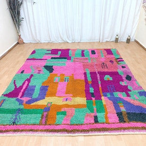 K&ouml;nnte beinhalten: Ein bunter, abstrakter Teppich mit geometrischen Formen in den Farben Rosa, Gr&uuml;n, Orange, Lila und Blau. Der Teppich hat einen Fransenrand.