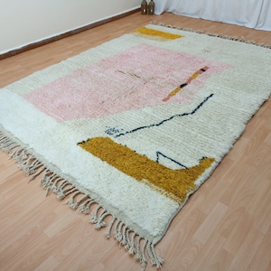 May include: A white, cream, pink, and yellow geometric patterned rug with fringe on the edges. The rug has a modern abstract design.