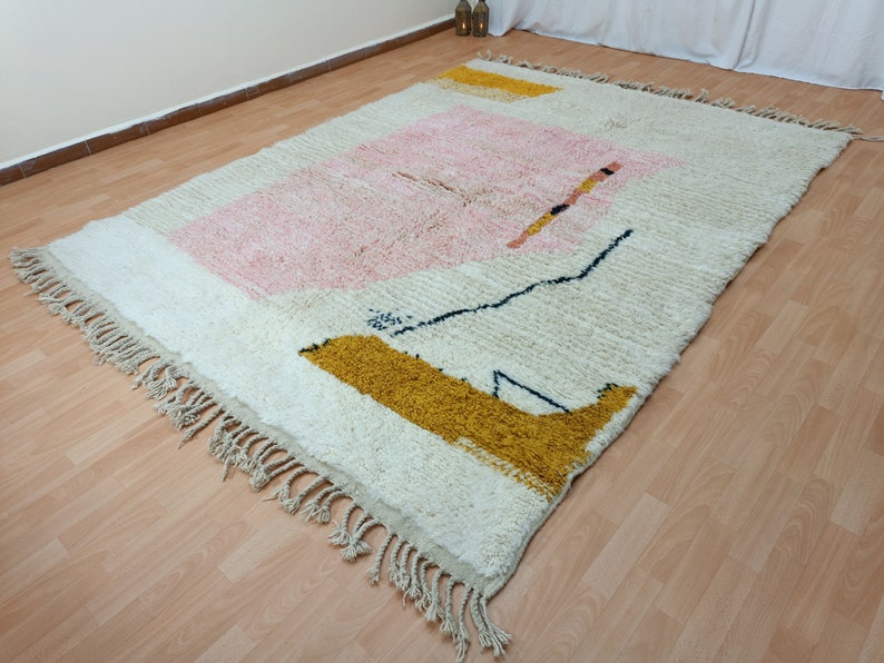May include: A white, cream, pink, and yellow geometric patterned rug with fringe on the edges. The rug has a modern abstract design.
