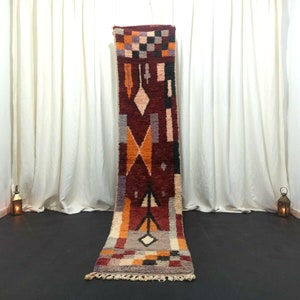 May include: A long, narrow, red, orange, white, and black patterned rug with a fringe on the ends. The rug is laid out on a black floor in front of a white curtain.